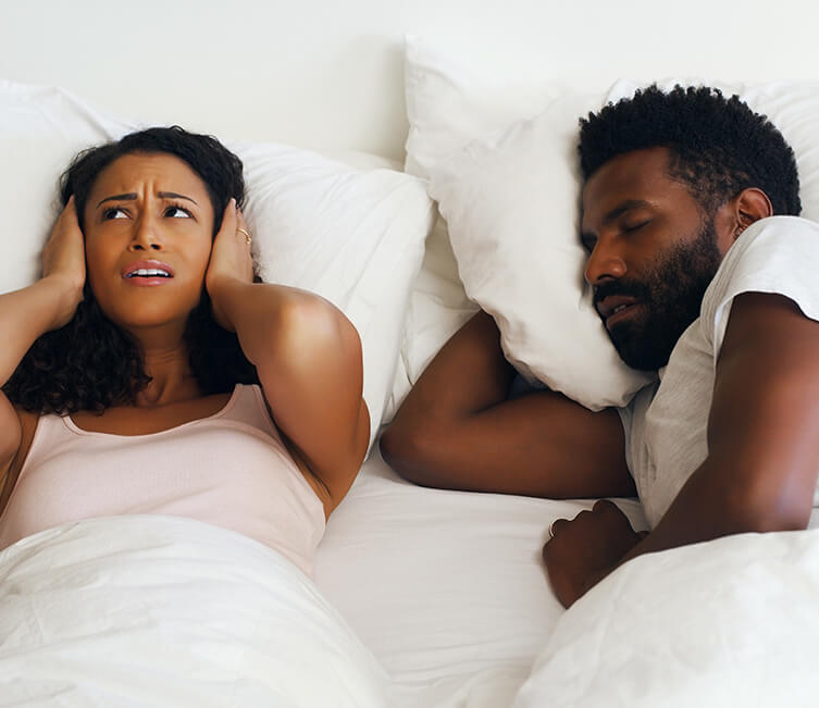 woman covering her ears while laying next to her snoring husband